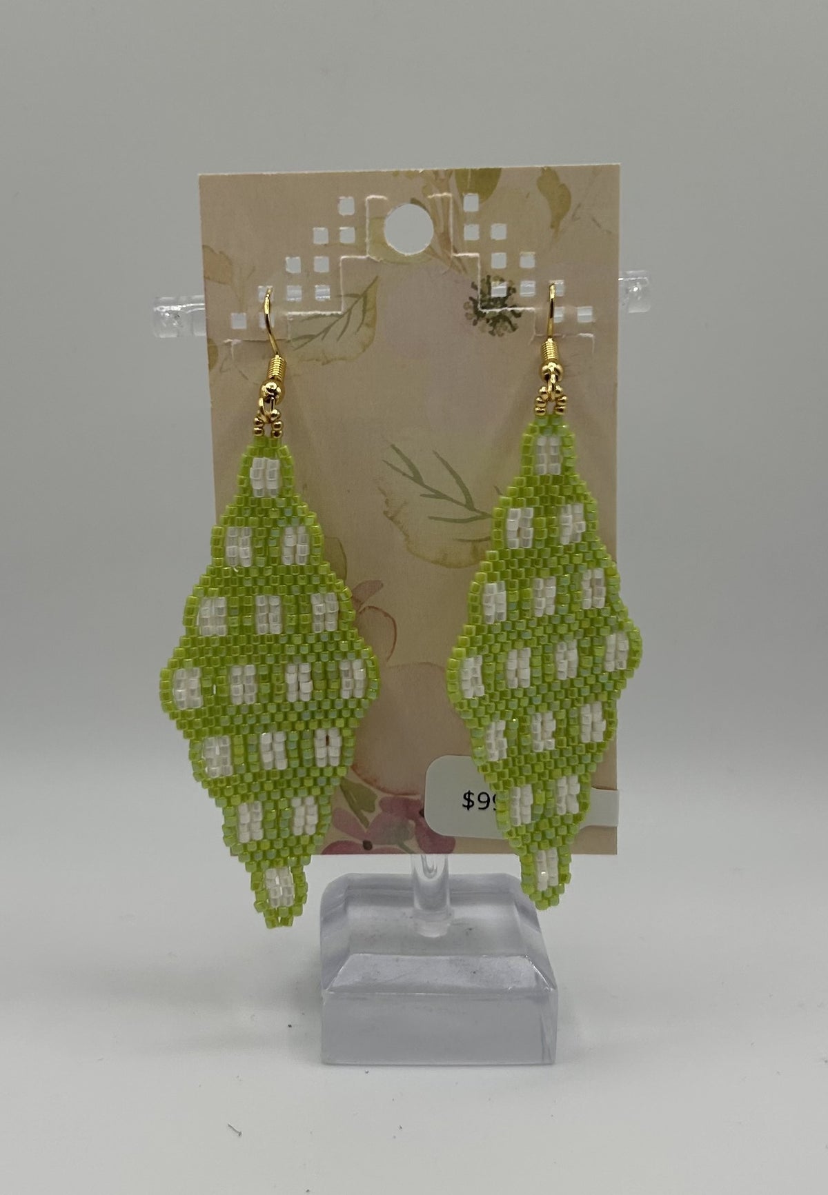 A pair of handmade beaded dangle earrings in light green and white, hanging from gold-toned hooks on a display card mounted on a clear stand.