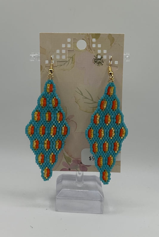 Handmade beaded dangle earrings with a turquoise blue base, orange and yellow patterned details, and gold hooks, displayed on a clear stand in front of a decorative backing card.